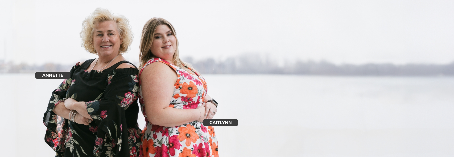 Two women, Annette and Caitlynn, stand back-to-back smiling in front of a blurred wintery lakeside background. Annette wears a black floral dress, while Caitlynn wears a white dress with bright orange and pink flowers.